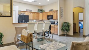 A modern kitchen with wooden cabinets, a refrigerator, and a microwave. There are chairs and a glass table in the dining area. - Las Cruces Vacation Rental Near Trails & Golf! (Las Cruces)