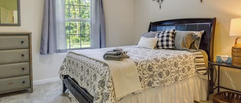 A bedroom with a bed, a dresser, a window with curtains, a wall-mounted lamp, and a wall decor piece.
