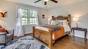 A bedroom with a wooden bed, two bedside lamps, a window with curtains, a chair, and a ceiling fan. - Hendersonville Haven w/ Fire Pit, Grill, Deck! (Hendersonville)