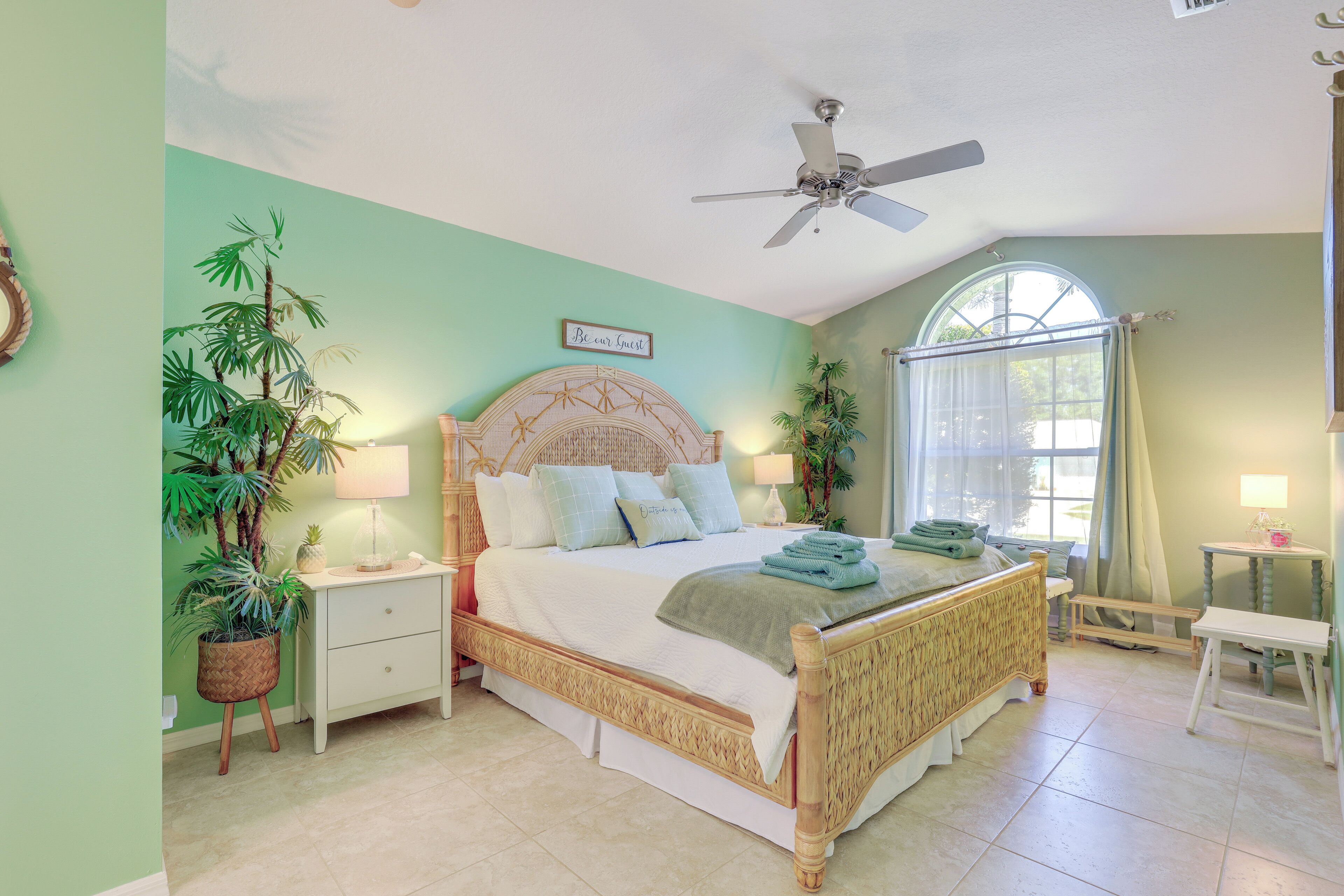 A bedroom with a bed, nightstands, a ceiling fan, a window with curtains, and potted plants.