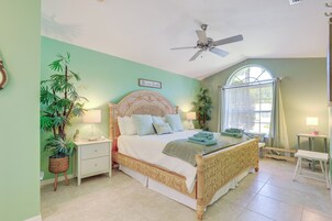 Une chambre à coucher comprenant un lit, des tables de chevet, un ventilateur de plafond, une fenêtre avec des rideaux et des plantes en pot.