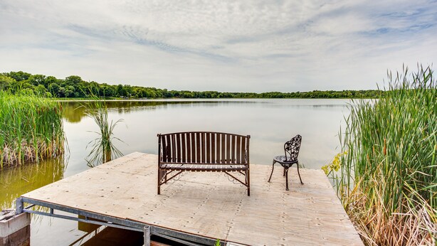 Un muelle de madera con un banco y una silla a orillas de un lago tranquilo, rodeado de follaje.