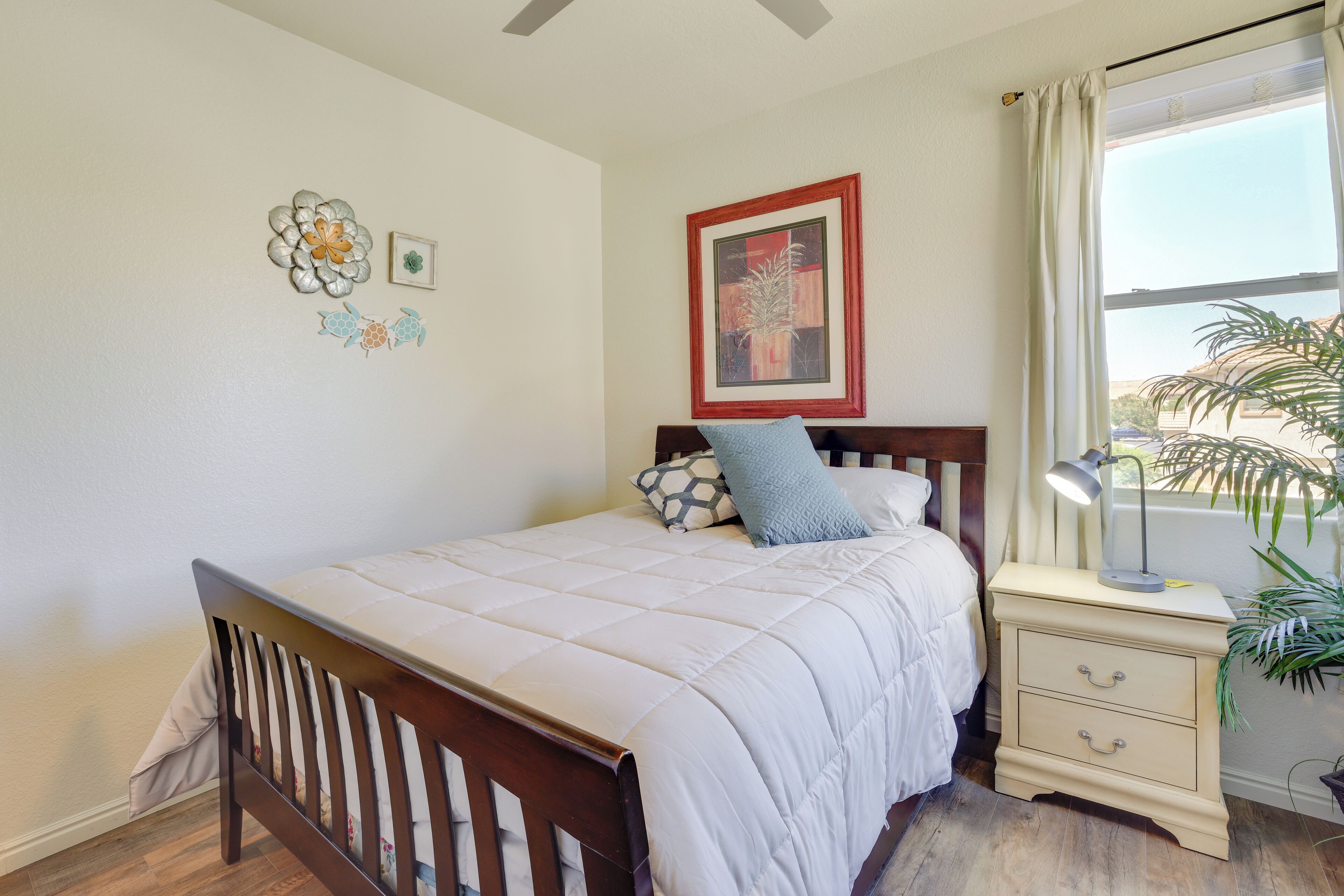 A bedroom with a wooden bed, a nightstand with drawers, a lamp, a window with curtains, and wall decorations.