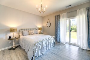 A bedroom with a large bed, two bedside tables with lamps, a chandelier, a sliding glass door with curtains, and a wall-mounted mirror. - Beaufort Getaway w/ Pool Access, 2 Mi to Downtown! (Beaufort)