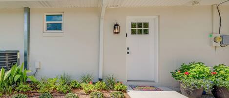A house with a white exterior, a front door, a small window, and a garden with various plants and flowers.