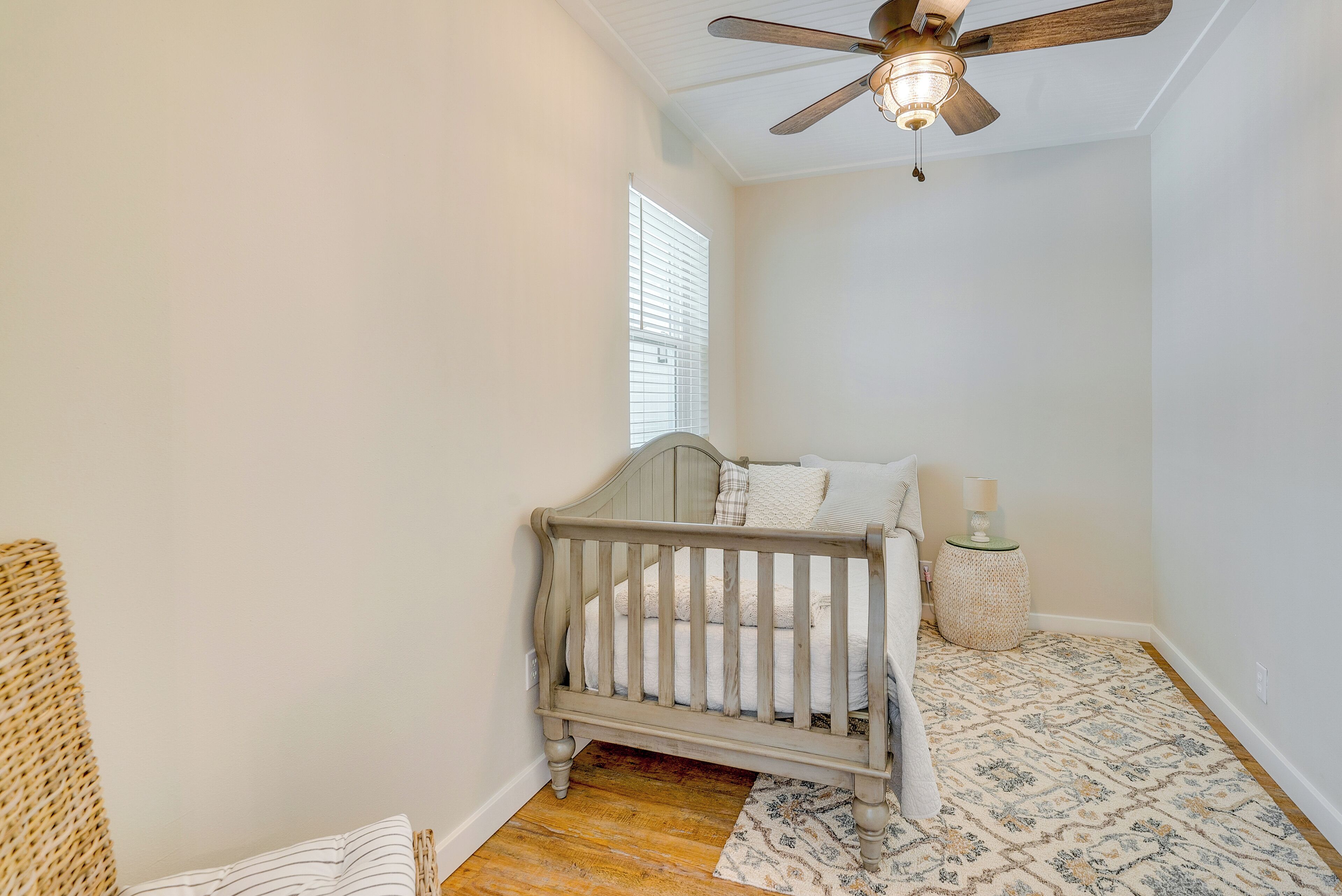 A small, well-lit room with a single bed, a ceiling fan, a window with blinds, and a patterned rug.