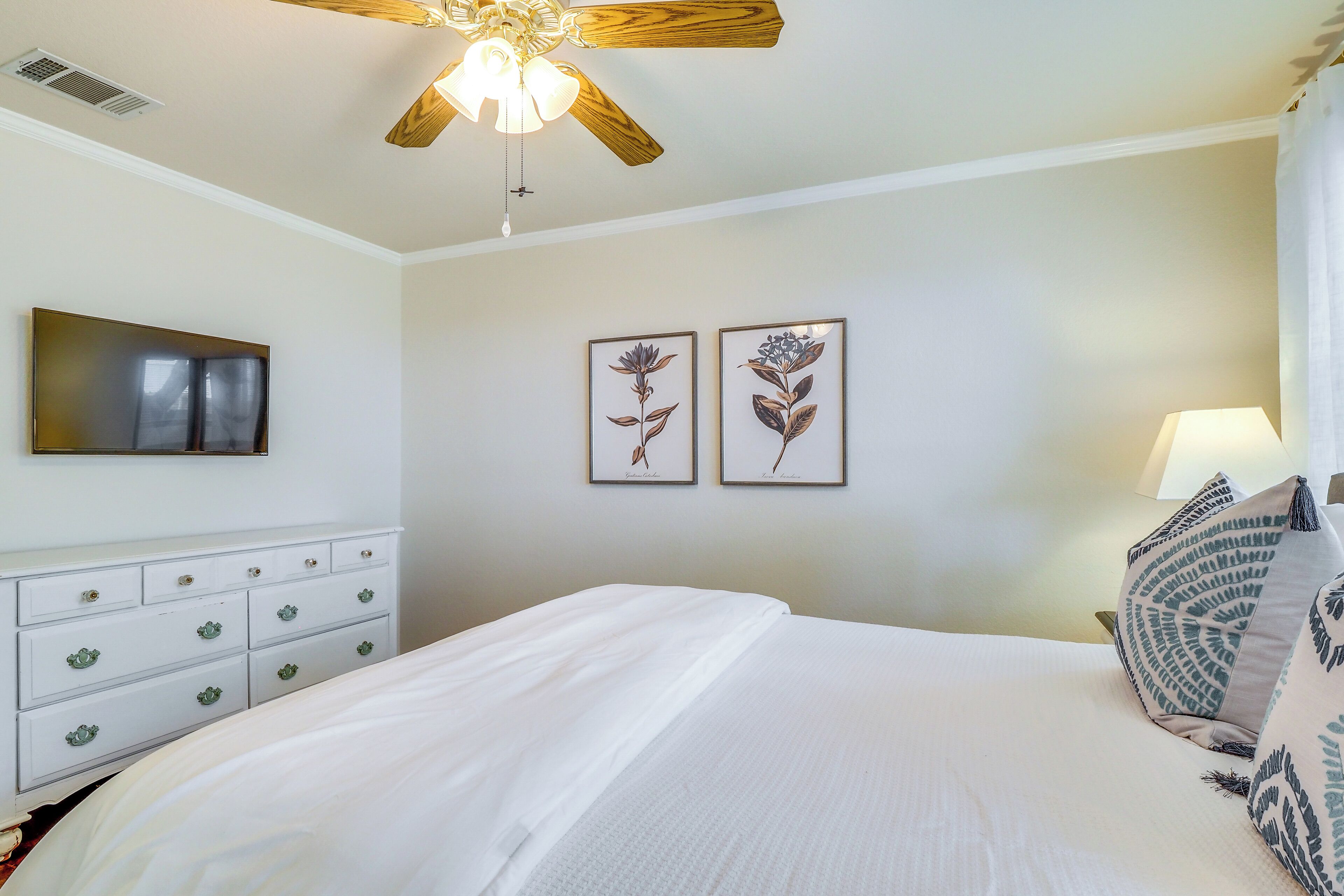 A bedroom with a bed, a ceiling fan, a dresser, and two framed botanical prints on the wall.