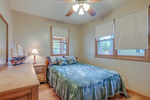 Quarto com cama, escrivaninha com modelo de navio, ventilador de teto e janela com persianas.
