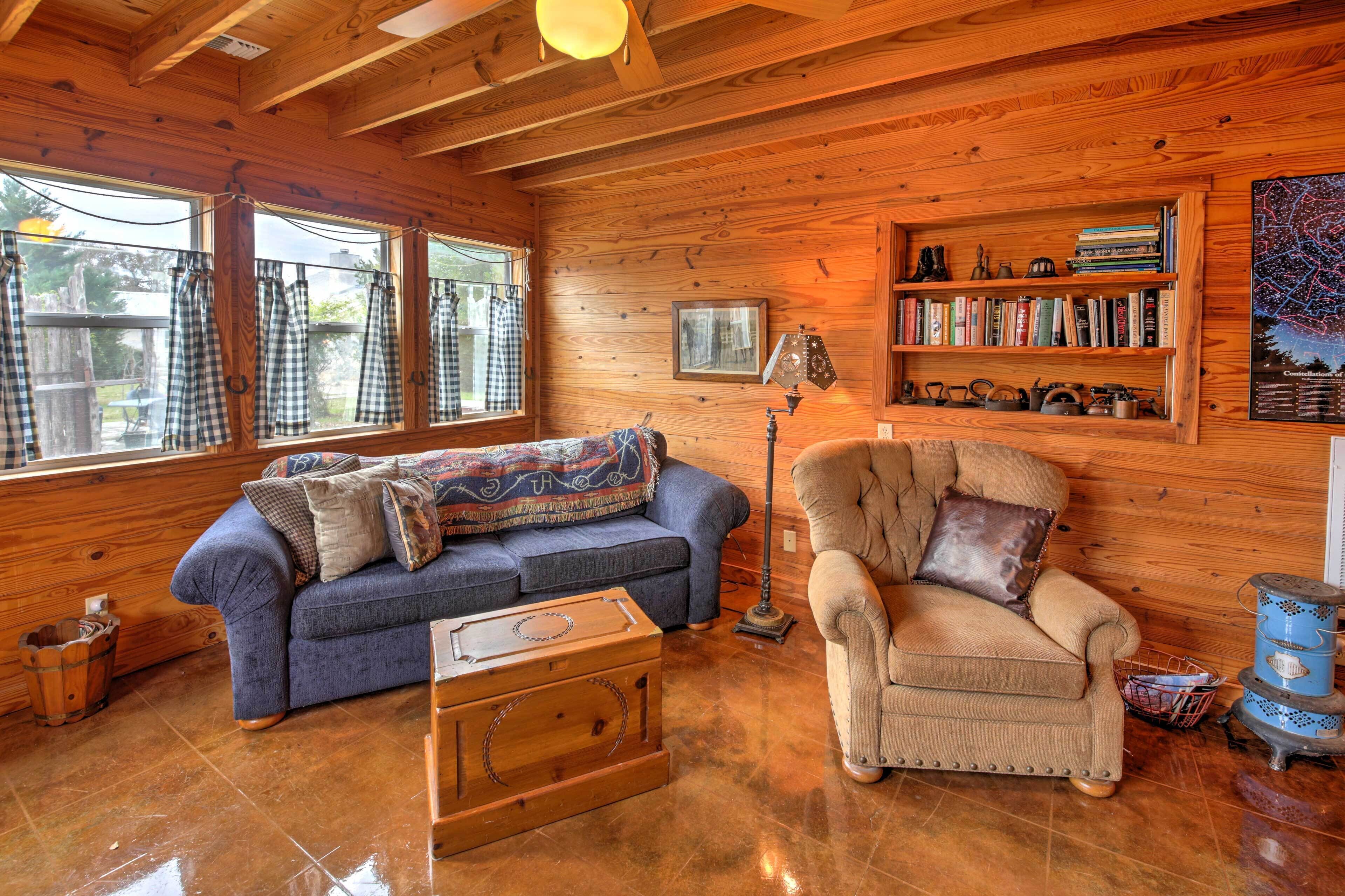 A cozy cabin interior with a wooden floor, a blue sofa, a beige armchair, a bookshelf, and a map on the wall.