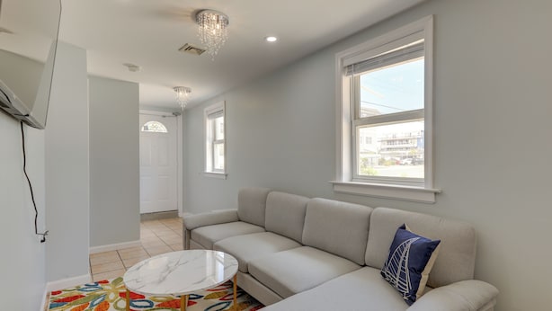 Ein modernes Wohnzimmer mit einem beigefarbenen Ecksofa, einem runden Couchtisch und einem farbenfrohen Teppich. Zwei Fenster sorgen für natürliches Licht und bieten einen Blick ins Freie.