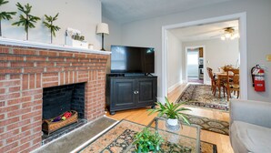 A living room with a brick fireplace, a flat-screen TV, a black entertainment unit, and a dining area with wooden furniture. - Pet-friendly Travelers Rest Home w/ Covered Patio (Travelers Rest)