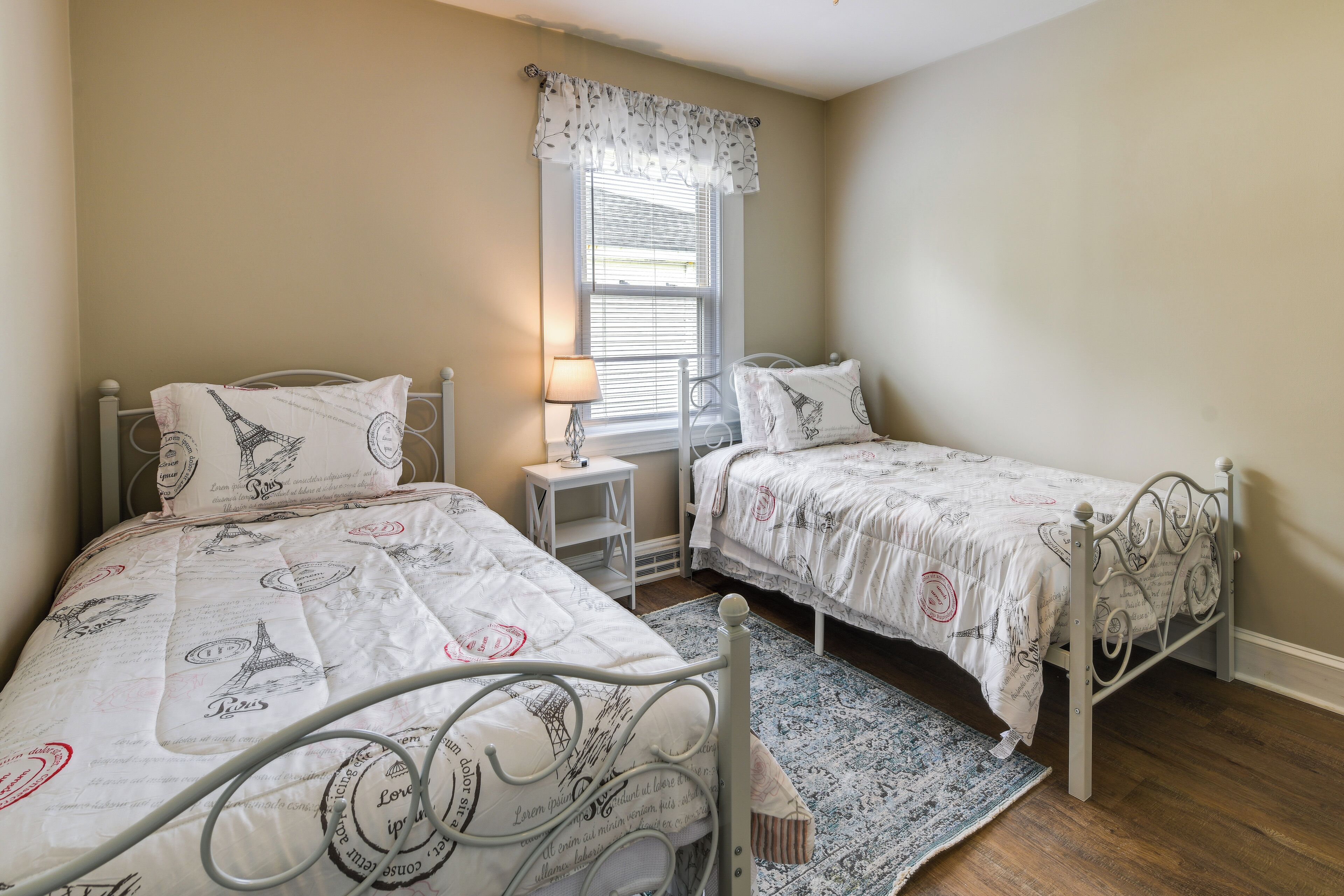 A bedroom with two beds, a window with blinds, a bedside table with a lamp, and a patterned rug.