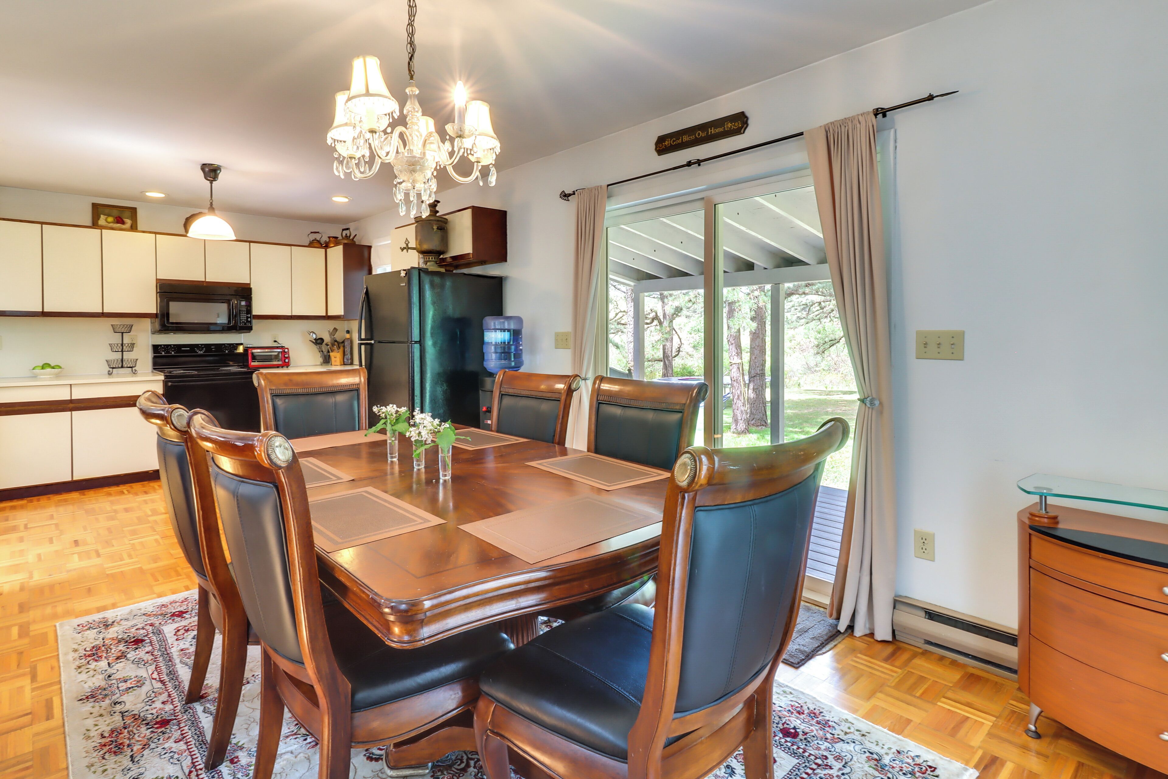 A modern kitchen with a dining area, featuring a wooden table, chairs, a chandelier, and a view through a glass door.
