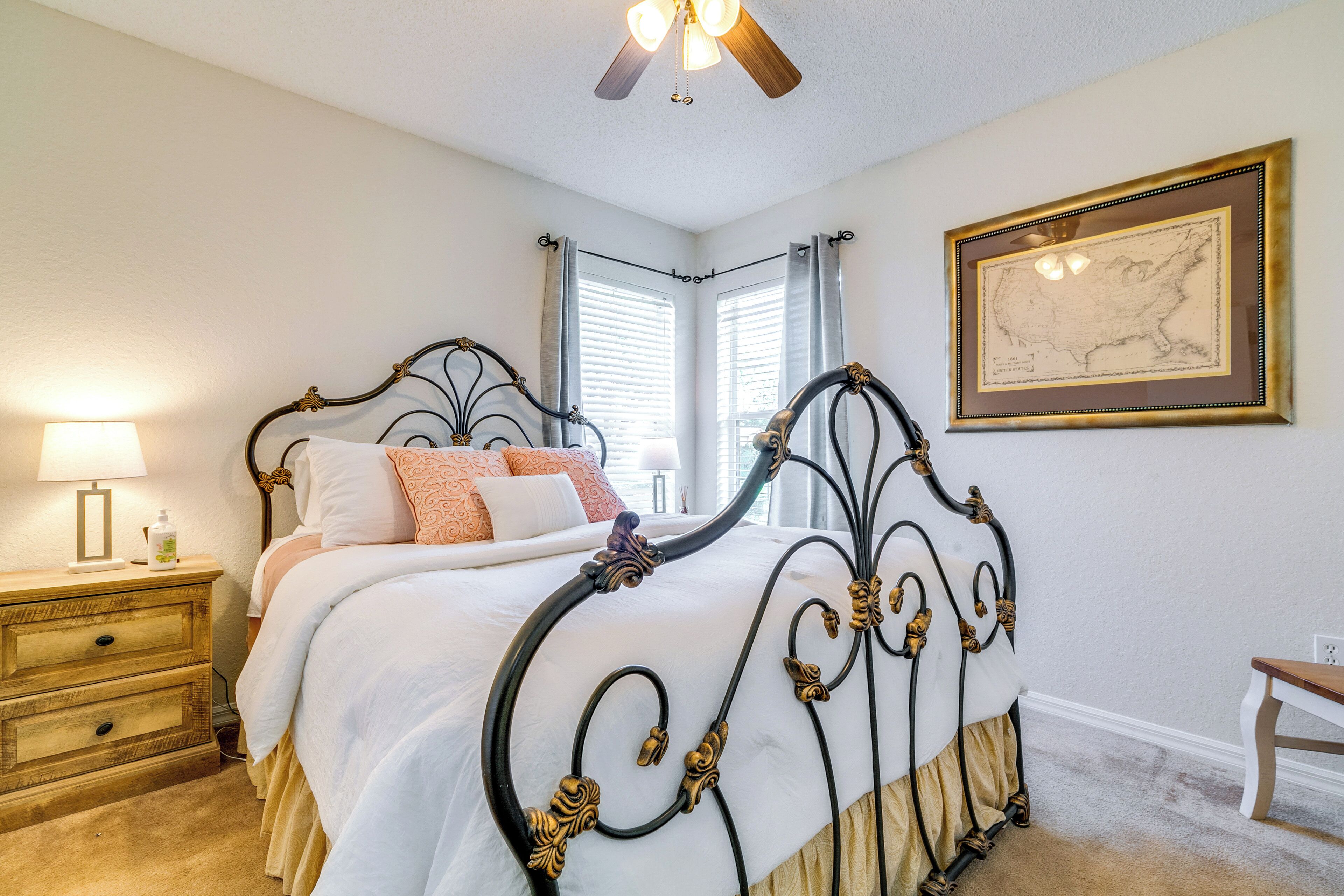 A neatly arranged bedroom with a classic bed, bedside tables, a nightstand, a ceiling fan, and a framed map on the wall.