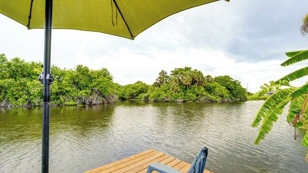 A wooden dock with a bench and a green umbrella, overlooking a calm river with dense greenery and trees.