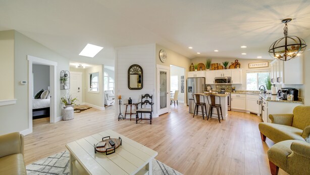 A spacious living room with a wooden floor, a dining area with a high table, and a kitchen with white cabinets and a farmhouse sign. - Coeur D'alene Vacation Rental ~ 4 Mi to the Lake! (Coeur d'Alene)