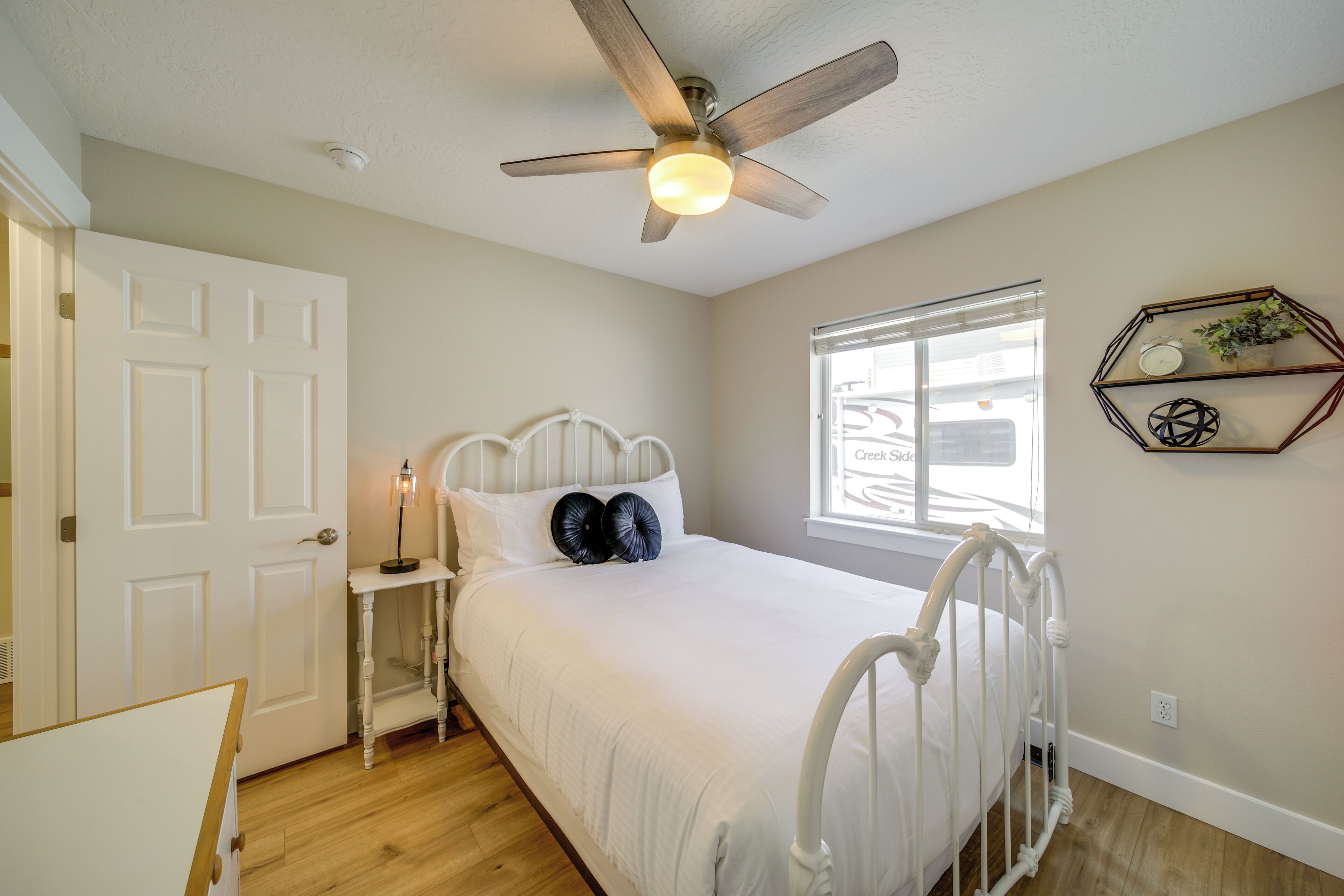 A bedroom with a white bed, a ceiling fan, a bedside table with a lamp, a window with a view, and a wall-mounted shelf with decorative items.
