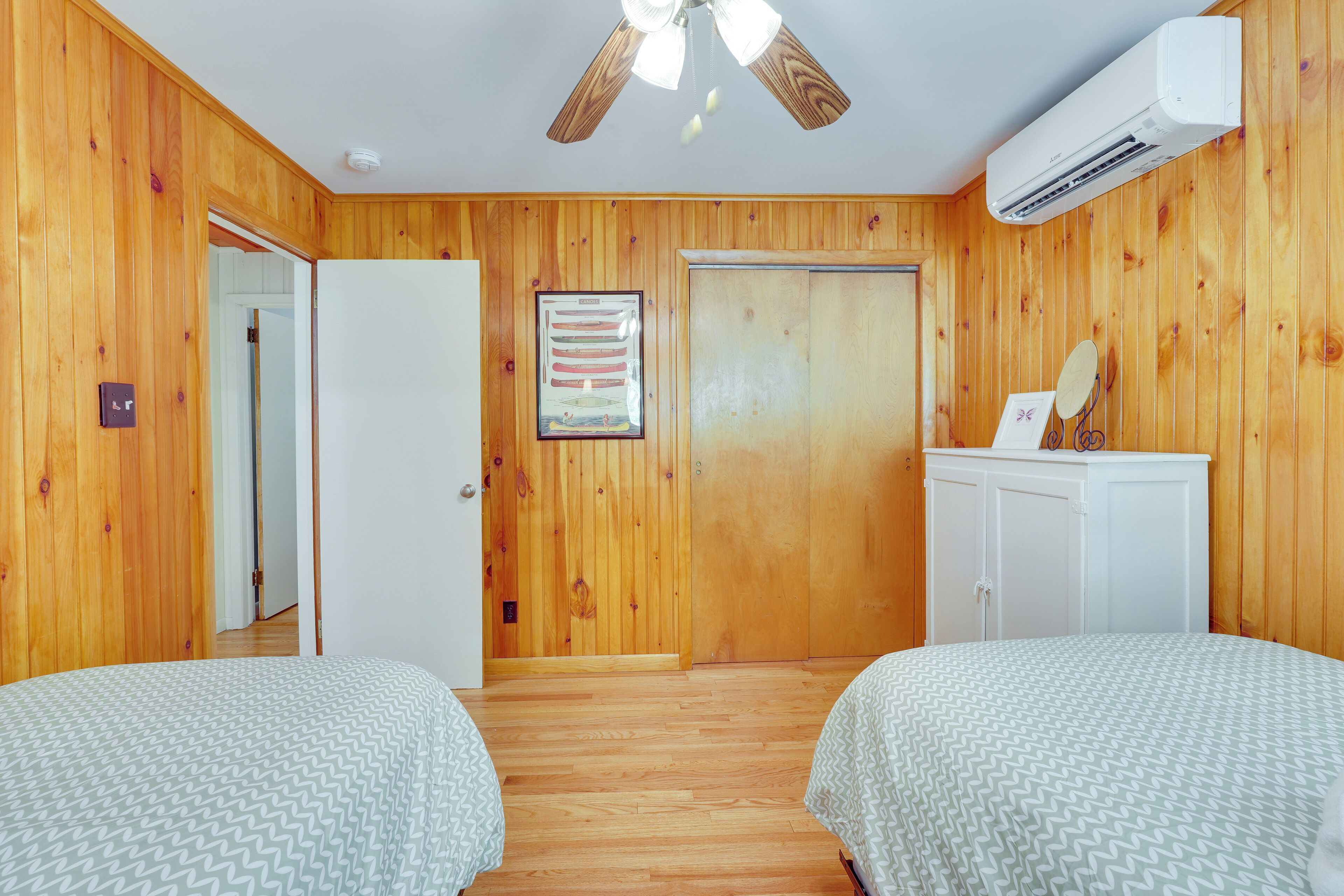 A room with wooden walls, a ceiling fan, an air conditioning unit, two beds with patterned bedding, a wardrobe, and a framed picture on the wall.