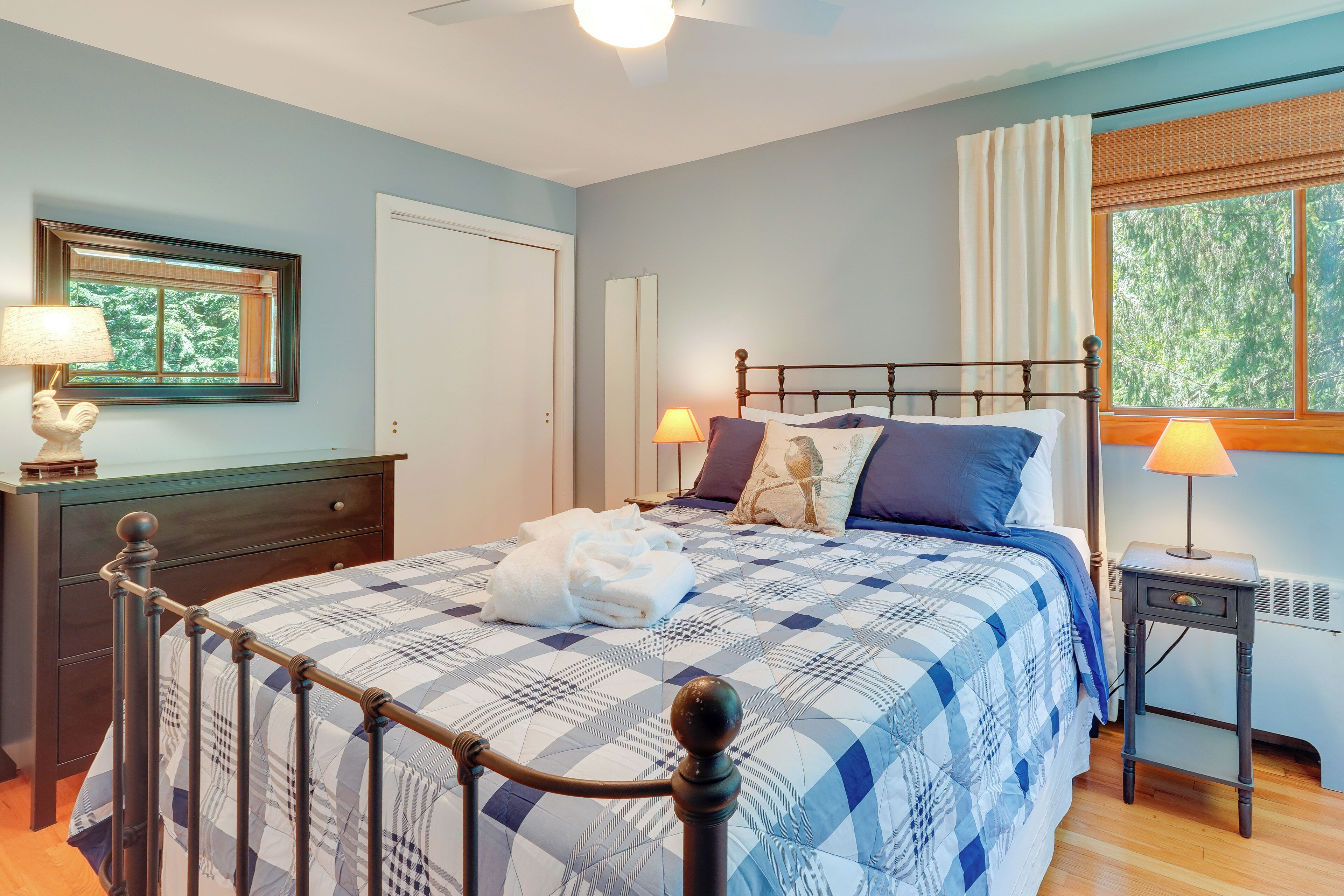 Una cama bien hecha con una colcha azul y blanca a cuadros, dos lámparas de noche y una ventana con cortinas.