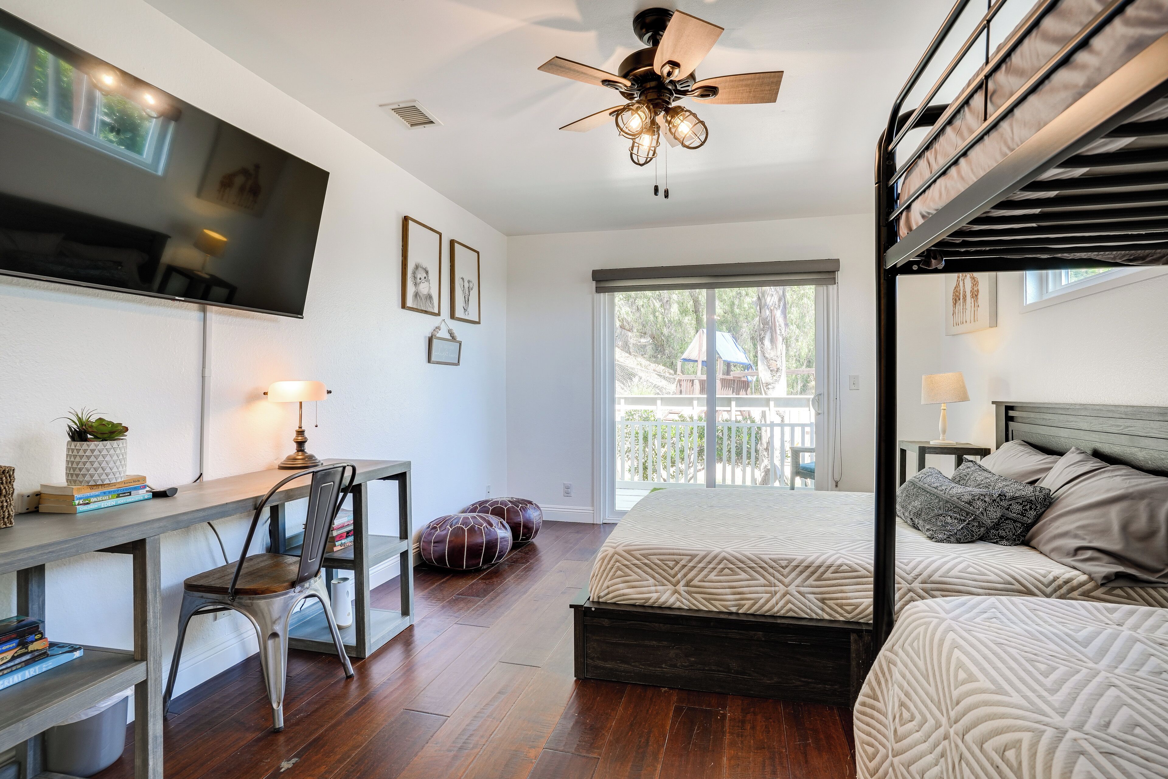 A bedroom with a bed, a desk, a chair, a ceiling fan, and a TV mounted on the wall.