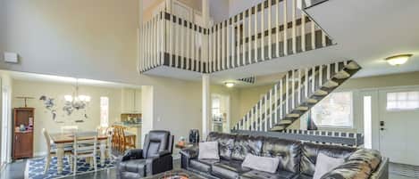 En rummelig stue med en stor sort lædersofa, et spisebord med stole og en trappe med stribet gelænder.