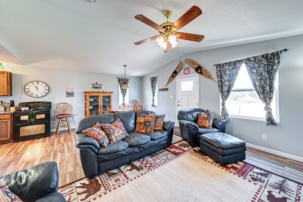 A spacious living room with a black leather sofa, a matching ottoman, and a patterned rug. A ceiling fan and a clock are visible on the wall.