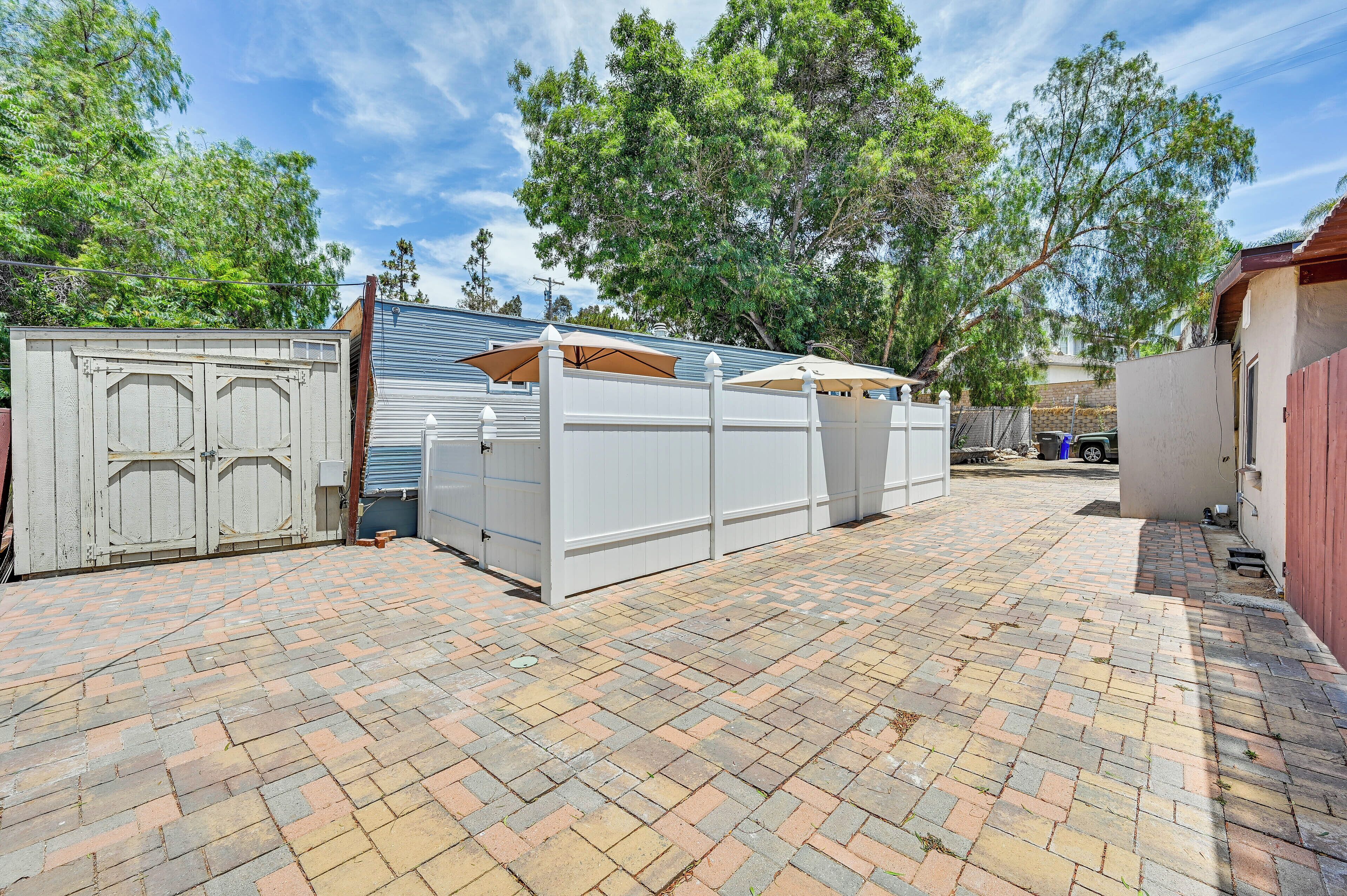 A paved backyard with a white fence, a covered patio area, and trees in the background.