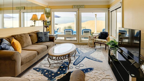 A cozy living room with a sofa, coffee table, and TV. Large windows offer a view of the desert. - Between the Dunes: Breezy Beachfront Oceano Home! (Oceano)
