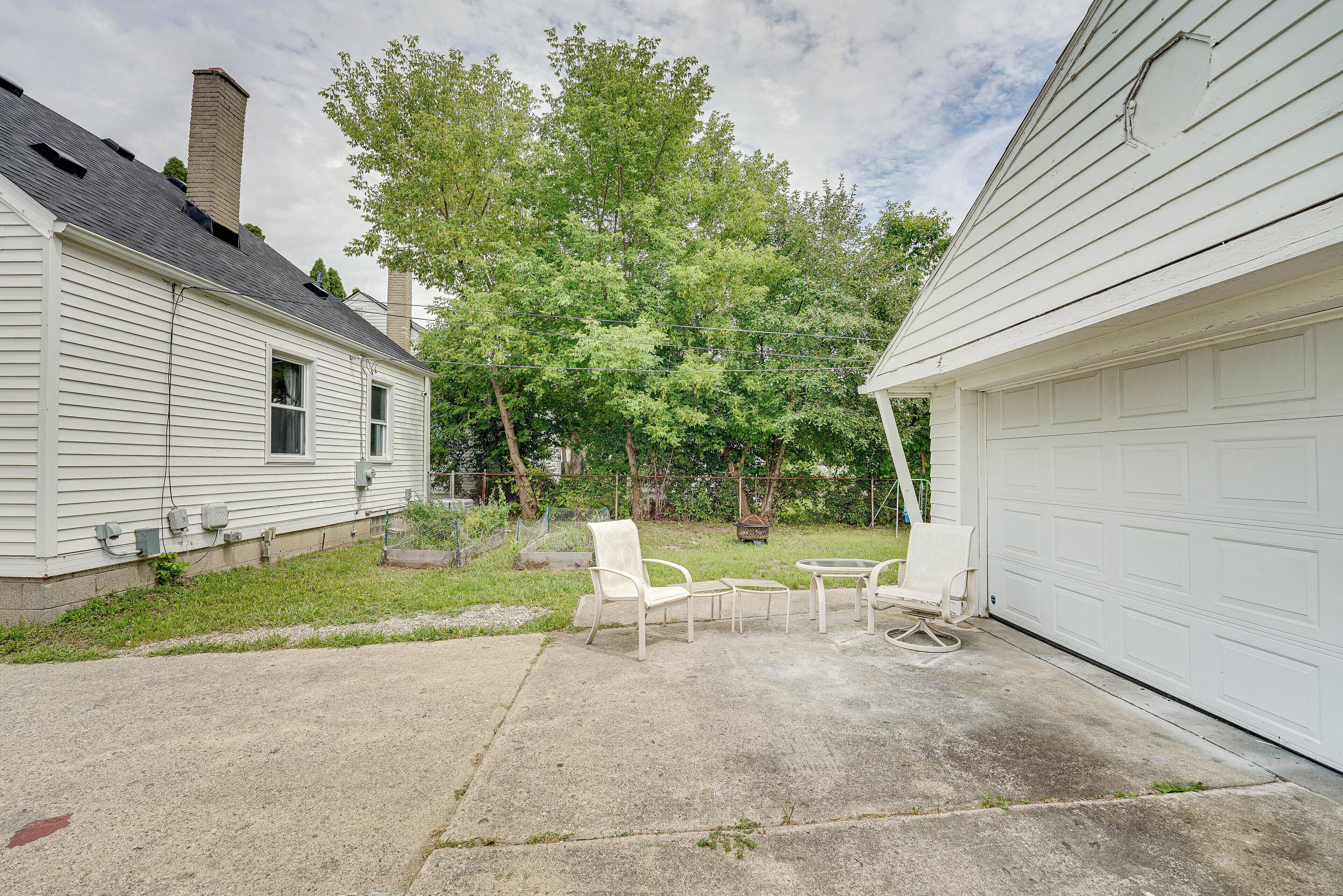 A residential backyard with a concrete patio, two white chairs, a small table, and a garage door.