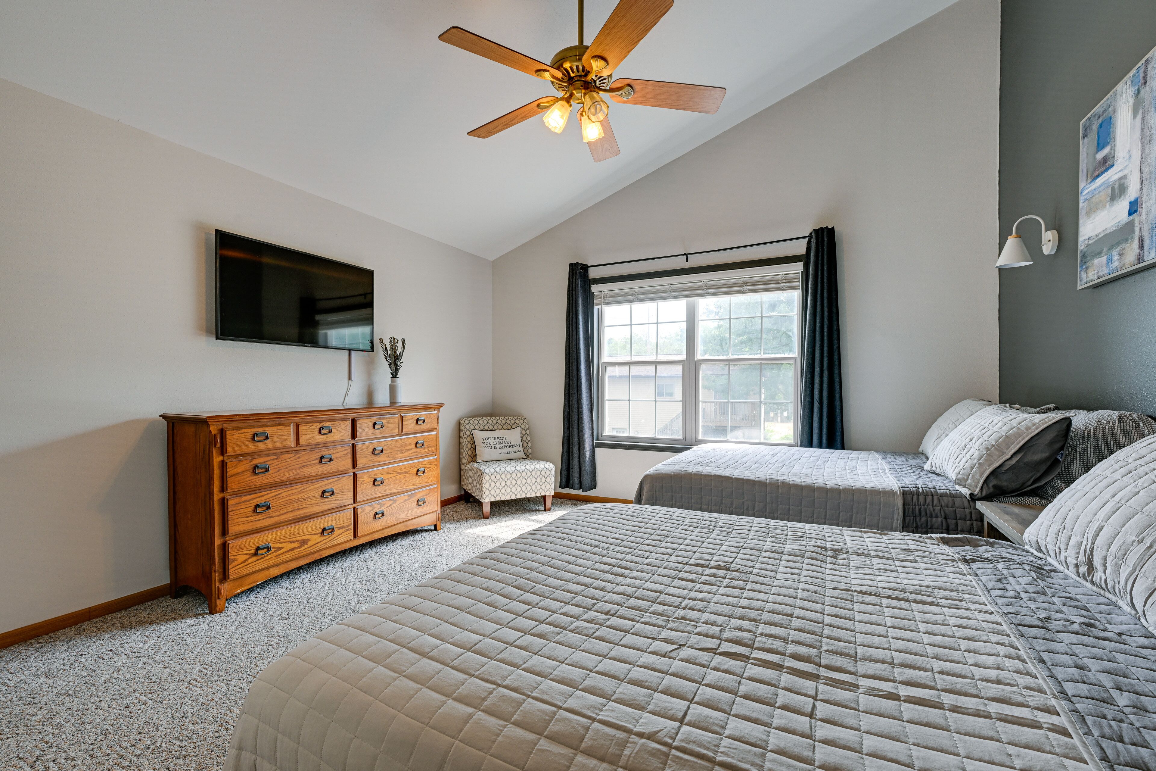 A bedroom with a large bed, a dresser, a chair, a ceiling fan, a TV, and a window with curtains.