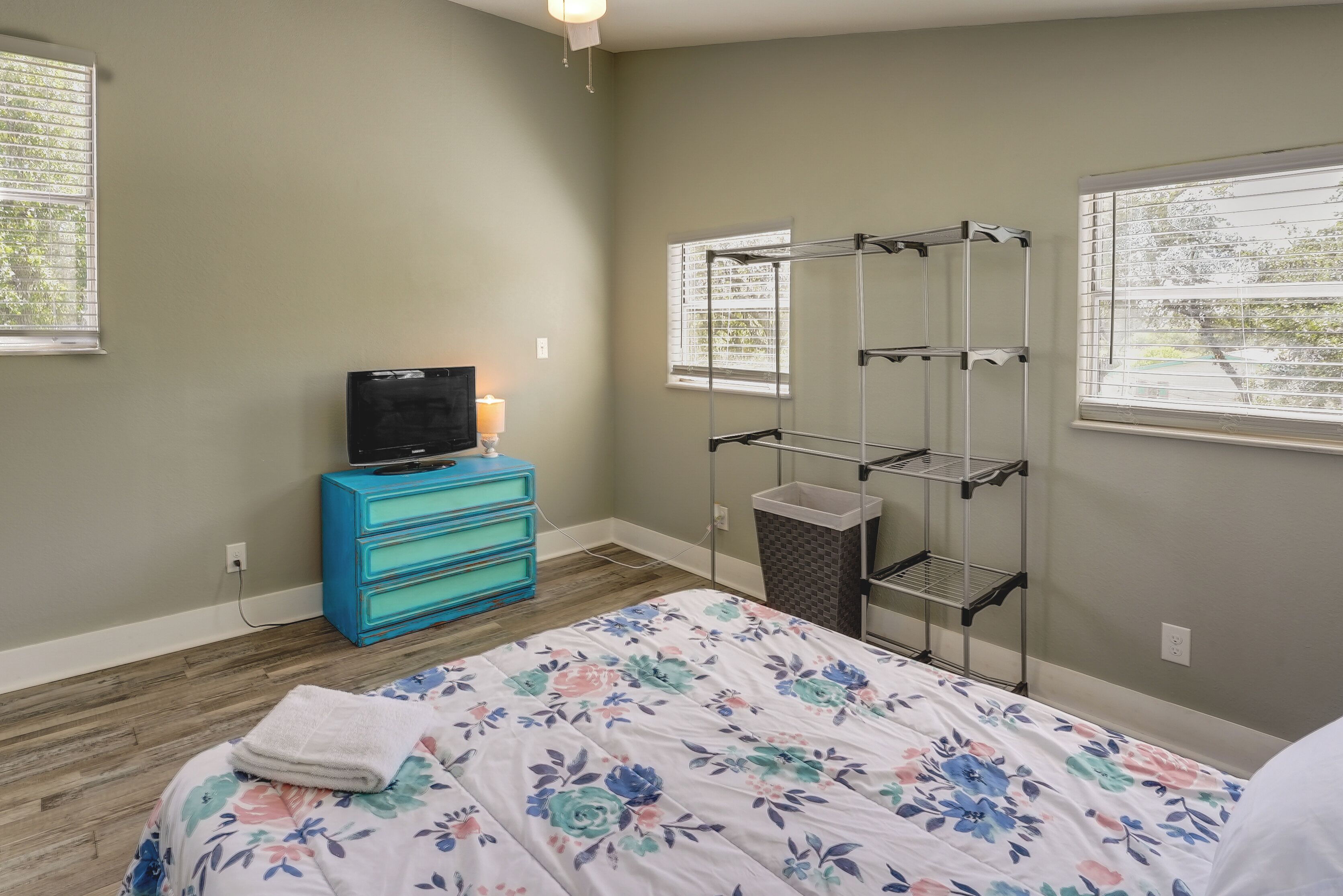 A bedroom with a bed, a blue dresser, a TV, a shelf unit, and a window.
