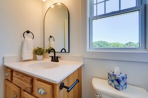 A bathroom with a white sink, wooden vanity, a mirror, a potted plant, a toilet with a tissue box, and a window with a view of greenery.