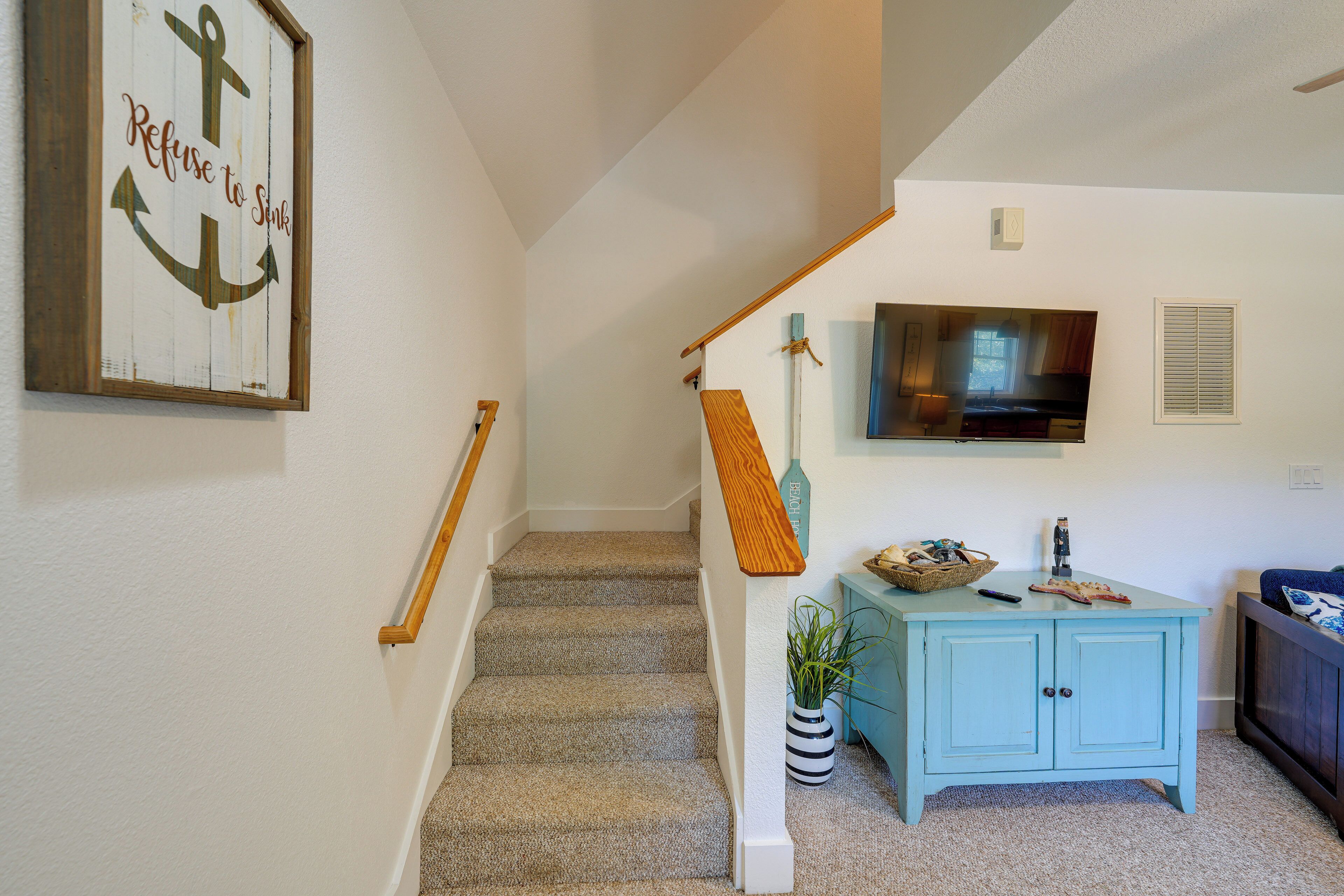 Eine Treppe mit Teppichstufen, einer Holzhandläufe, einem an der Wand montierten Fernseher, einem hellblauen Schrank und einem gerahmten Schild mit einem Anker.