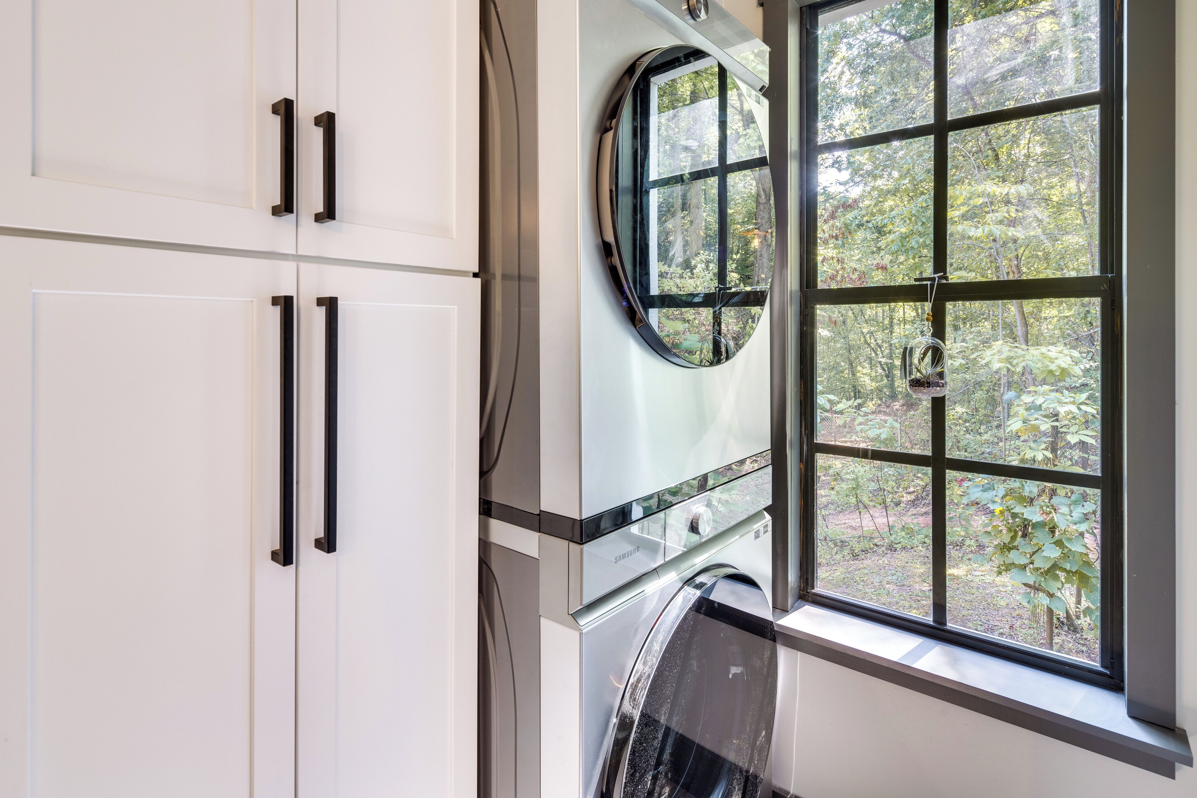 A modern laundry room with a built-in washer and dryer, a large window with a view of trees, and white cabinetry with black handles.