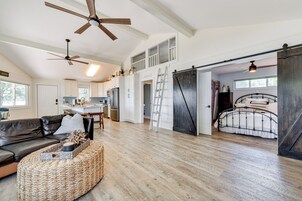 A spacious living area with a large sofa, a coffee table, and a ceiling fan. There is a kitchen area with cabinets and a refrigerator. A bed with a metal frame is visible in the background.