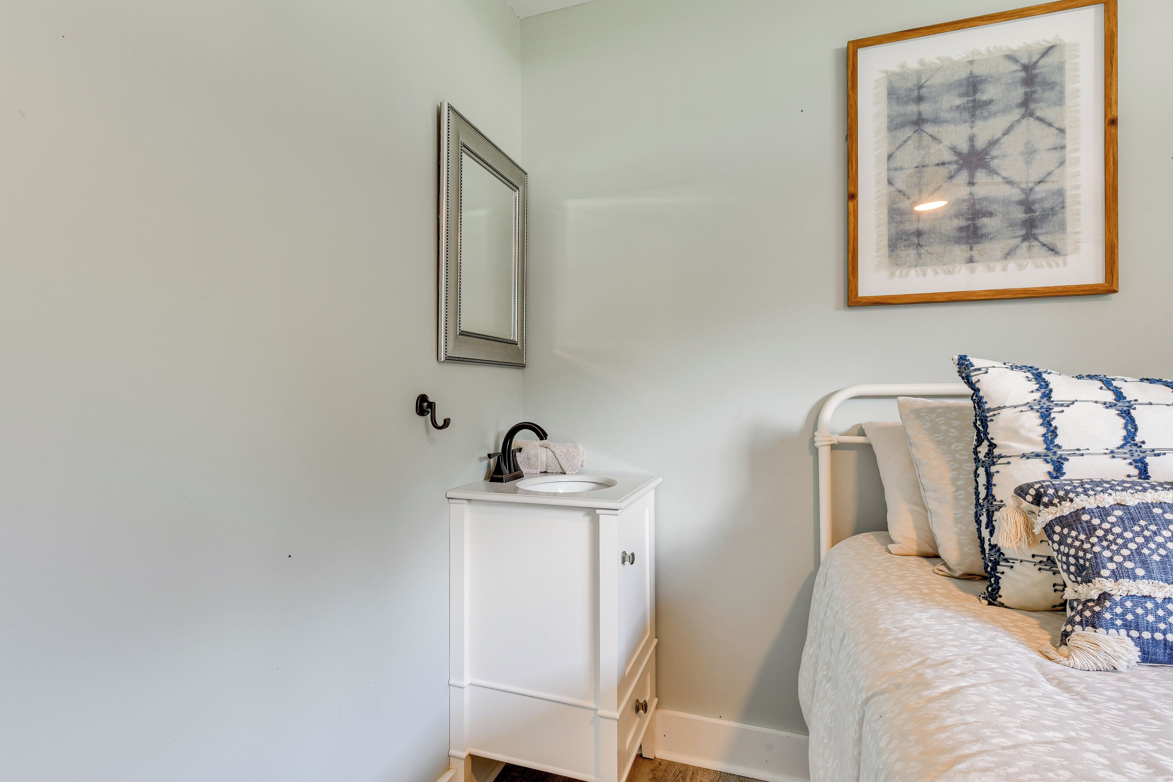 Una camera da letto con un letto, un comodino bianco con uno specchio, un piccolo mobile bianco con un lavandino e un'opera d'arte astratta incorniciata appesa al muro.