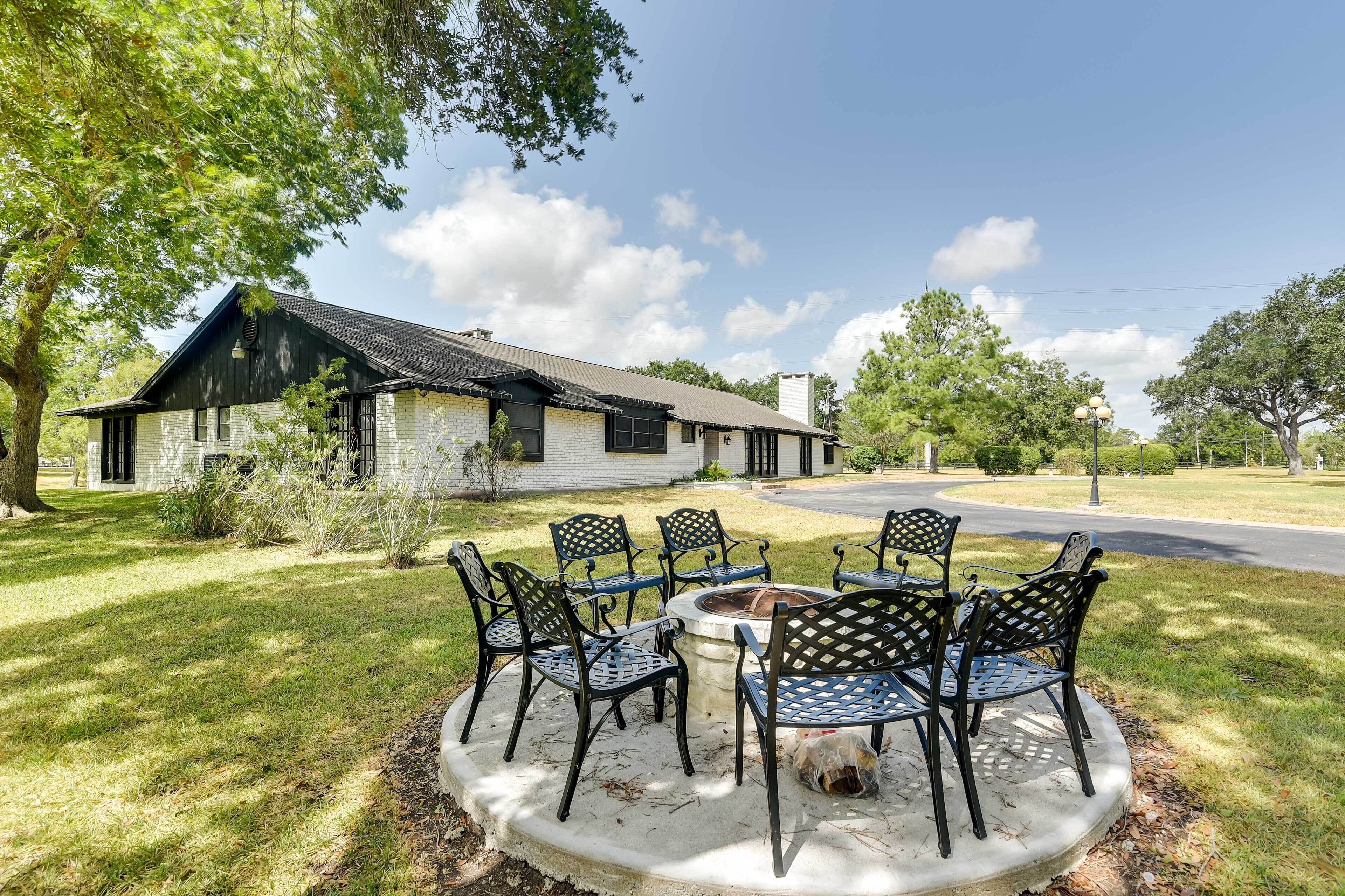 A stone patio with a fire pit and metal chairs, surrounded by a grassy lawn and trees.