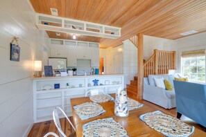 A cozy living room with a wooden ceiling, a white sofa, a blue armchair, and a dining table with crocheted placemats.