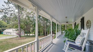 Eine überdachte Veranda mit weißen Geländern, eine Bank mit einem floralen Kissen und im Hintergrund ein weißes Gebäude sowie Bäume.
