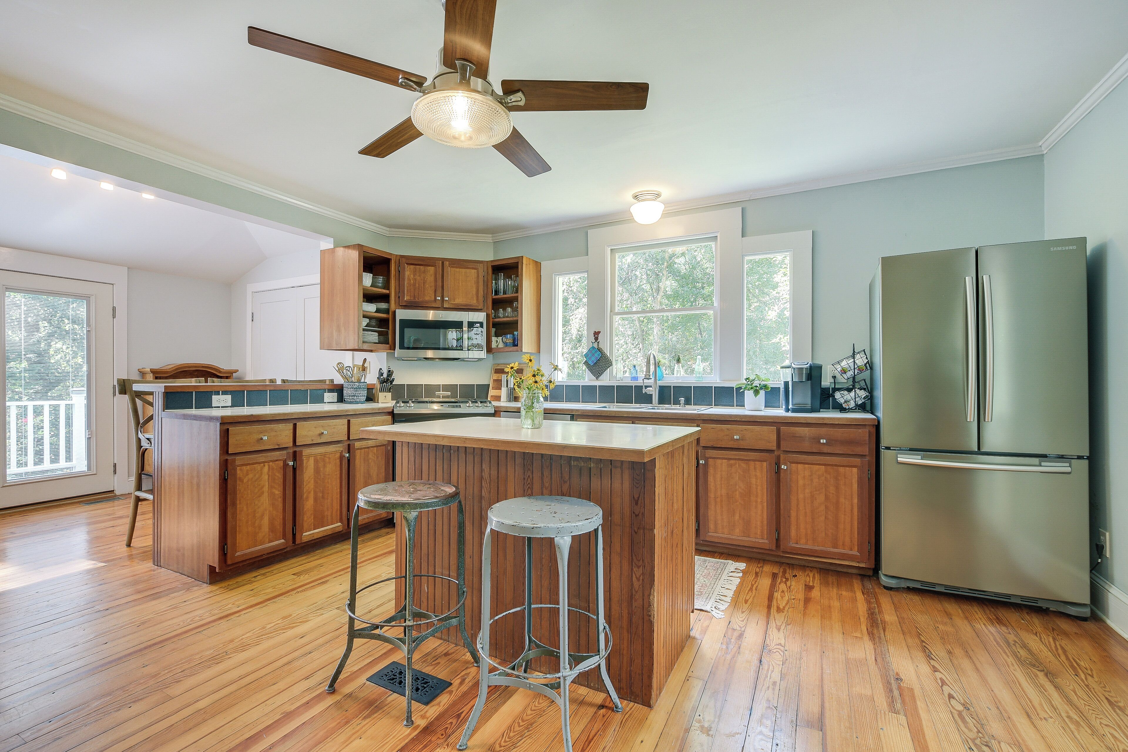A kitchen with wooden cabinets, a stainless steel refrigerator, a ceiling fan, and a large window.
