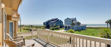 A wooden deck with chairs and a table, overlooking a beach with houses and a clear sky.