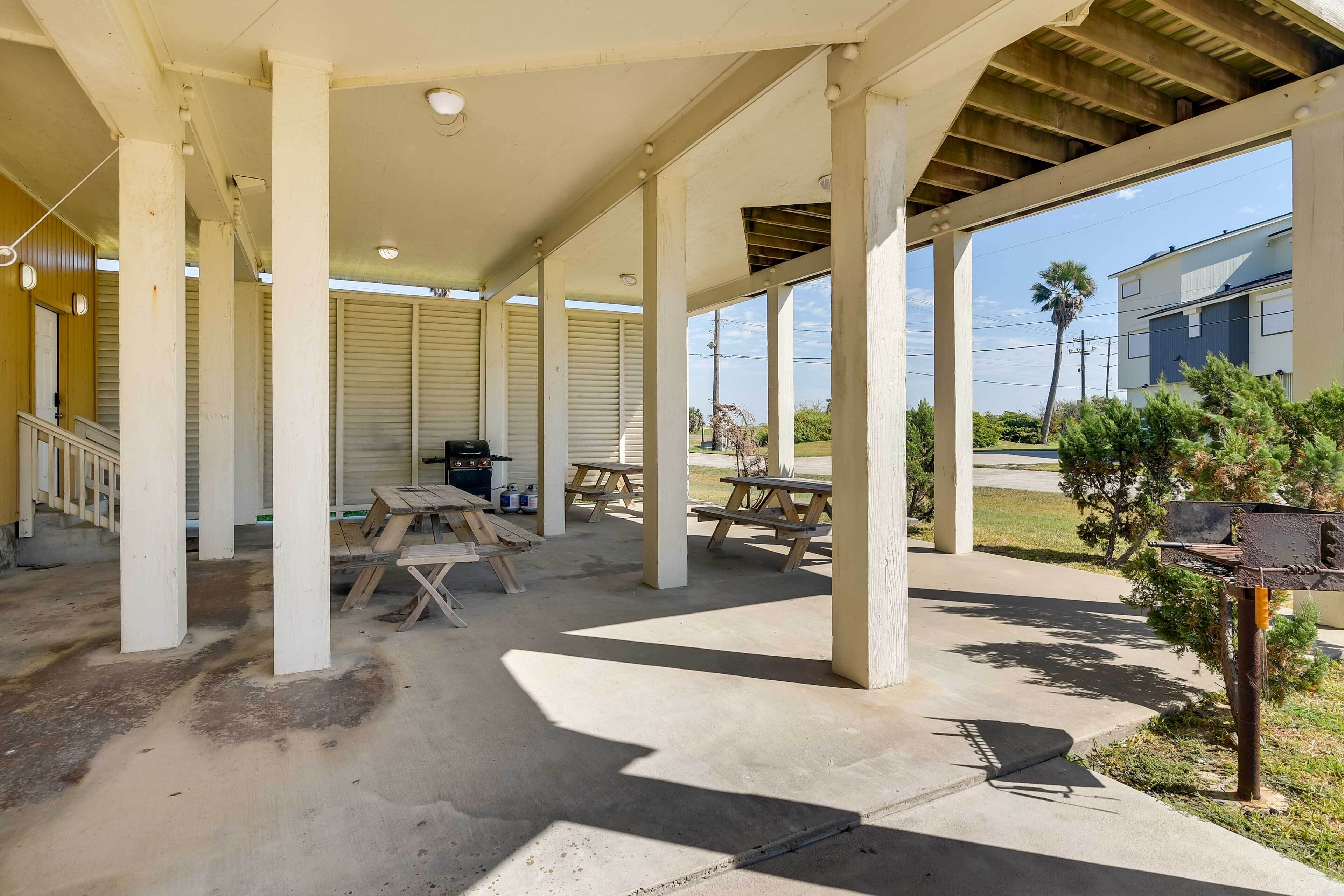 Un espacio al aire libre cubierto con mesas de picnic y una parrilla, columnas que sostienen el techo, y una vista de un edificio residencial al fondo.