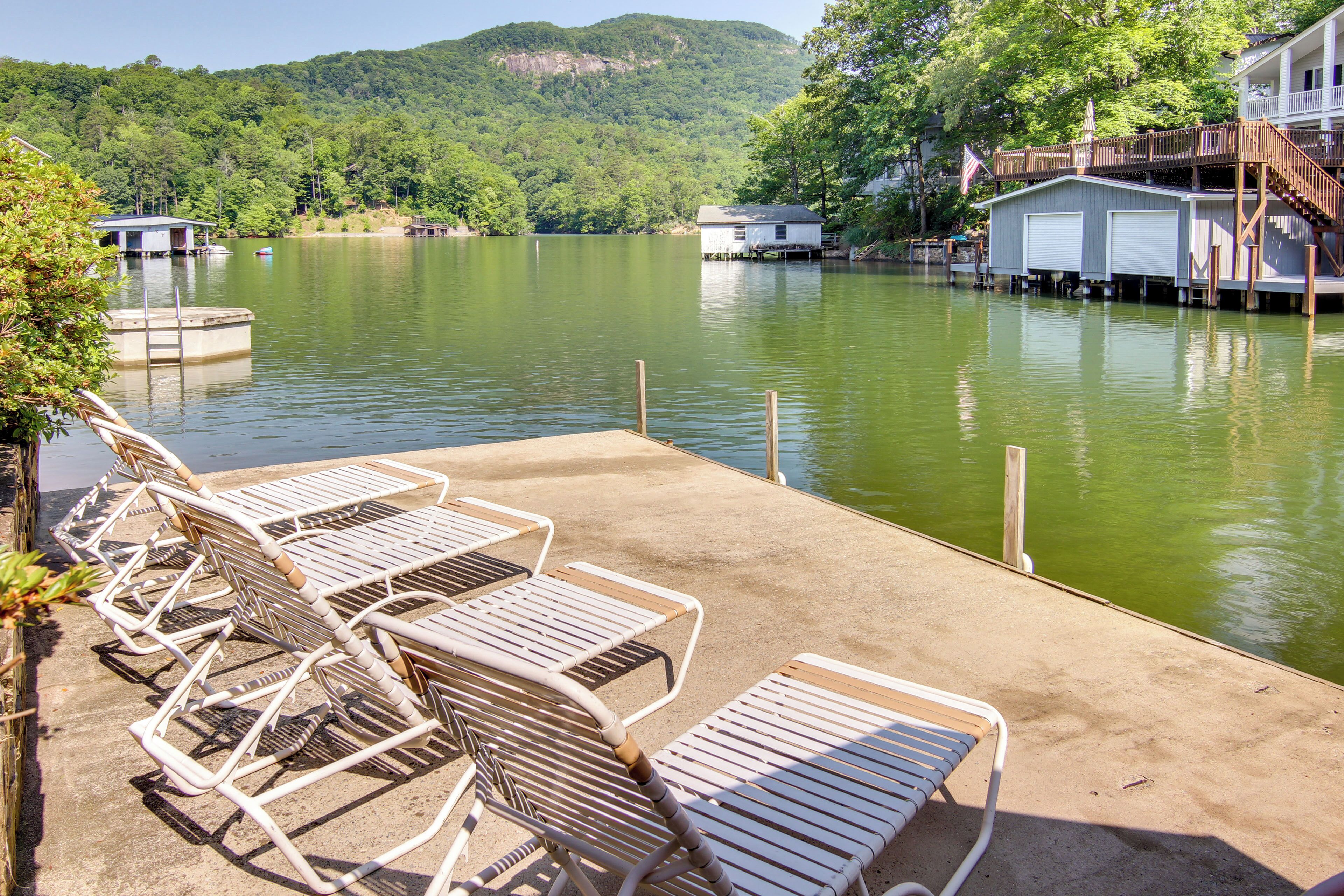 A lakeside dock with lounge chairs and a wooden deck overlooking a calm lake, surrounded by lush greenery and hills.
