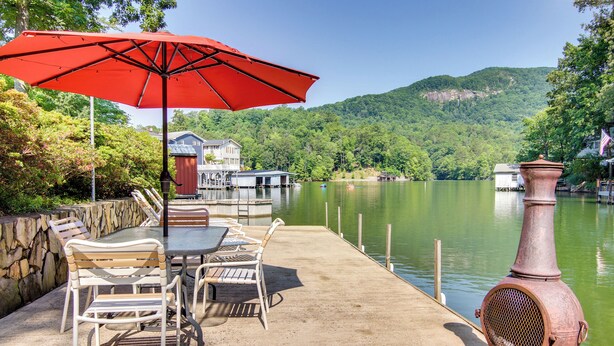 Une terrasse au bord du lac, avec une table et des chaises sous un parasol rouge, une cheminée rustique et une maison en arrière-plan.