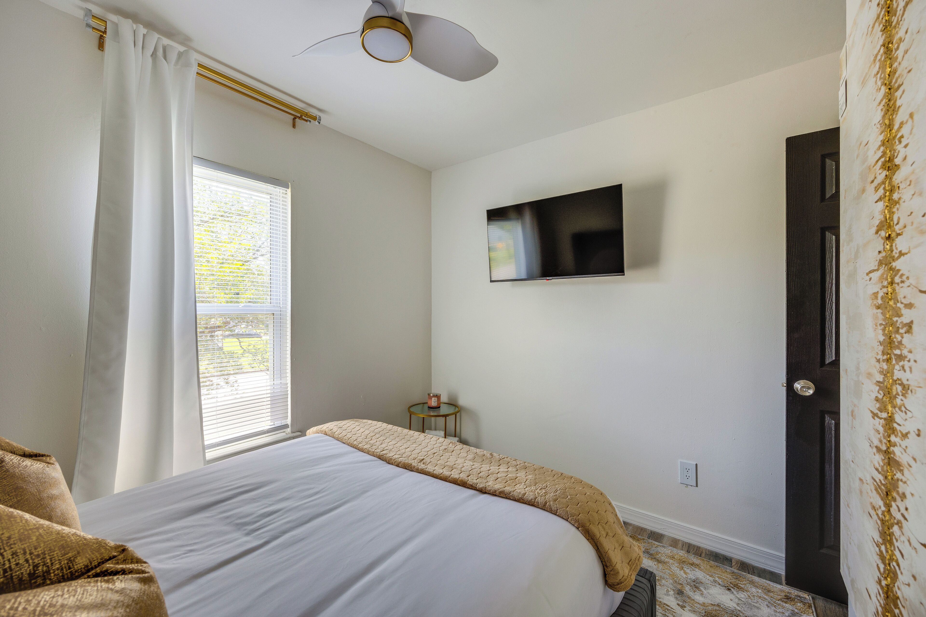 A bedroom with a bed, a ceiling fan, a TV mounted on the wall, a window with curtains, and a door with a decorative pattern.