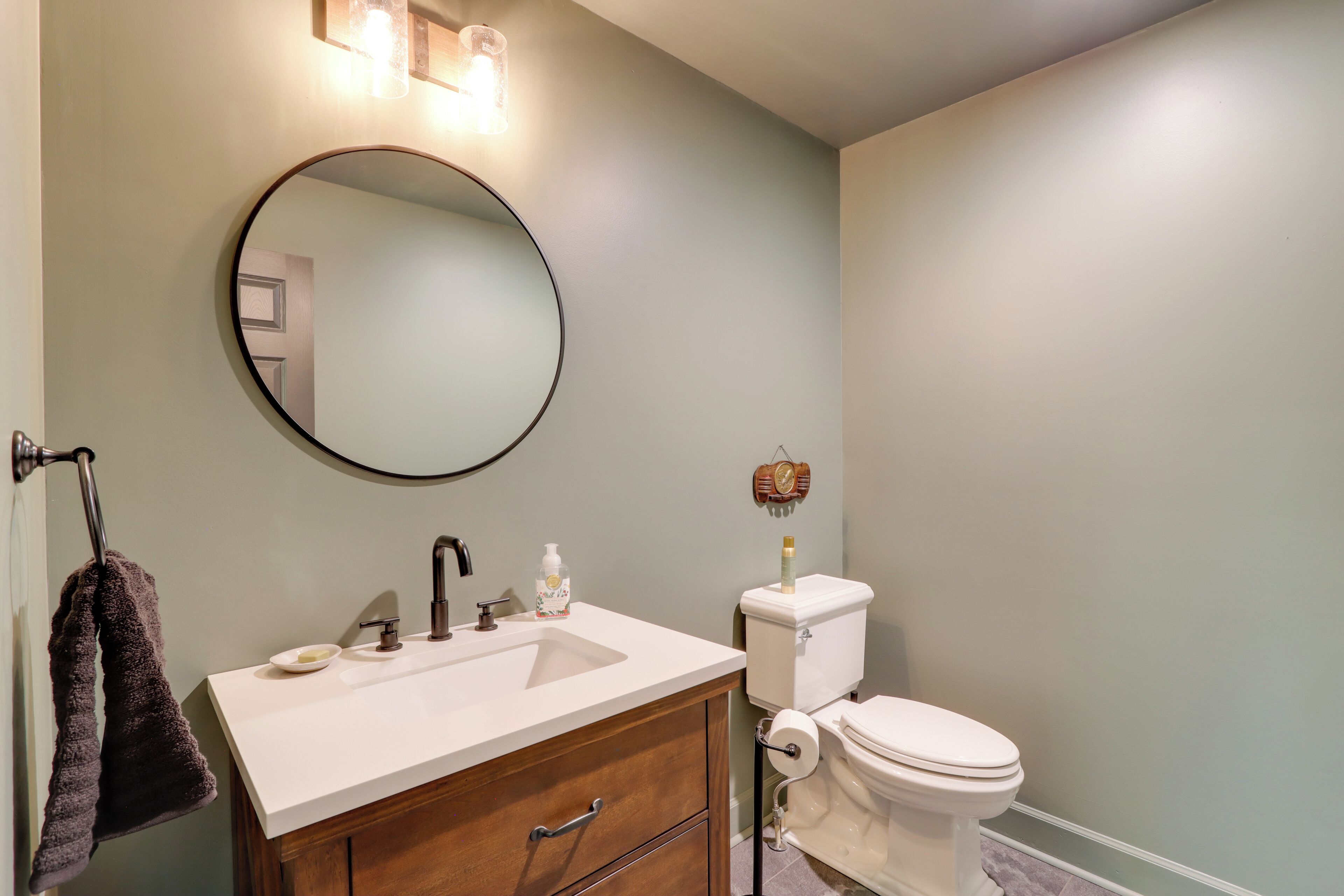 A bathroom with a round mirror, a white sink, a wooden cabinet, a toilet, and a towel hanging on a rack.