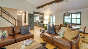 Een ruime woonkamer met een bruine leren bank, een salontafel en een eethoek met een houten tafel en stoelen. Een trap leidt naar een hoger gelegen niveau.