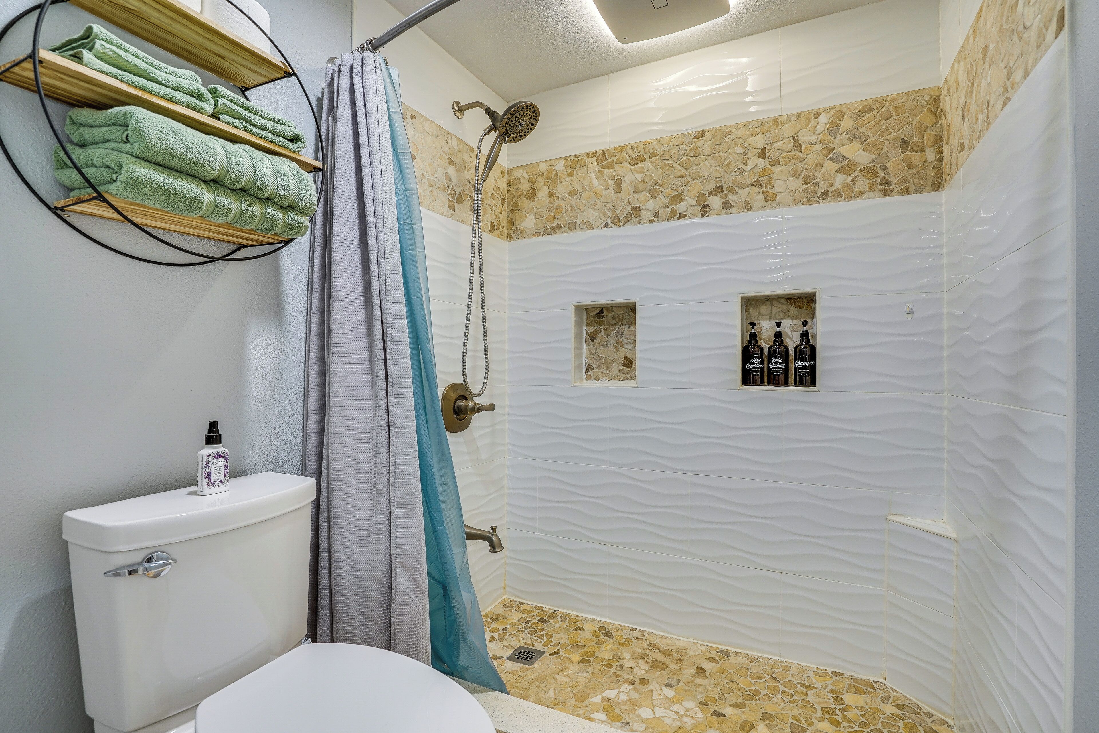 A bathroom with a toilet, a shower with a mosaic tile floor, and a towel rack with green towels.
