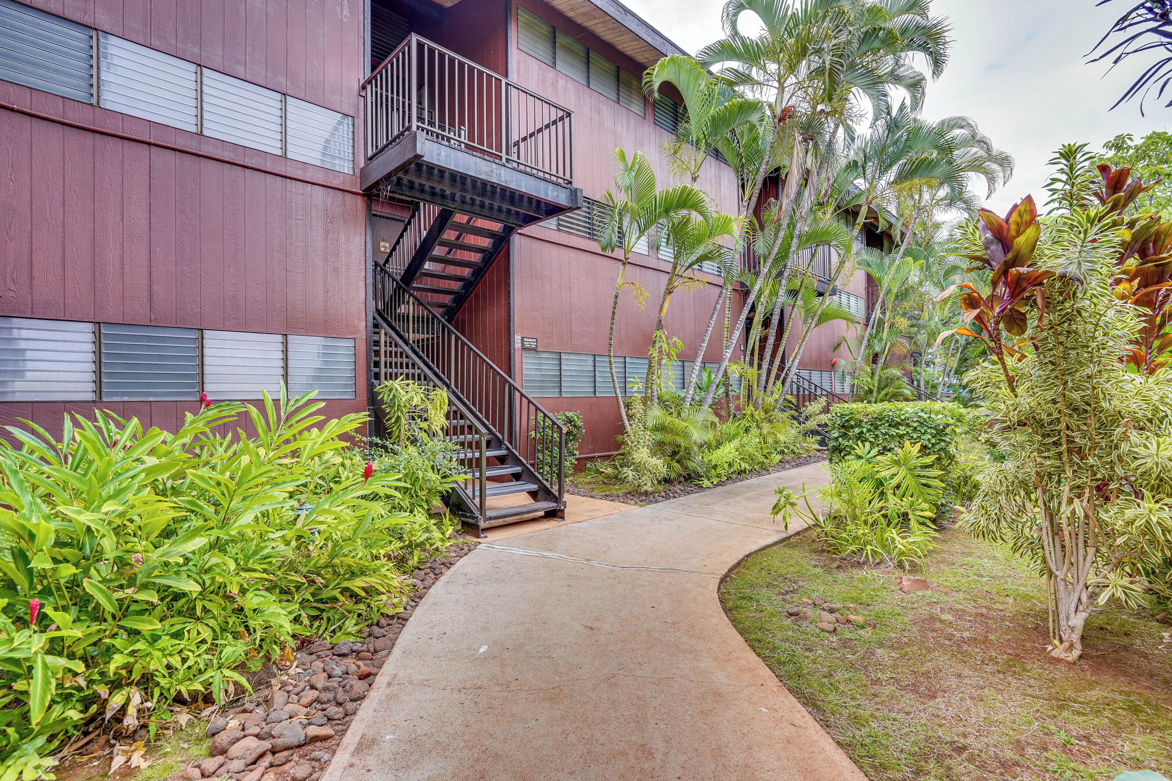 Un bâtiment à l’extérieur rouge, avec un chemin en béton et diverses plantes tropicales.