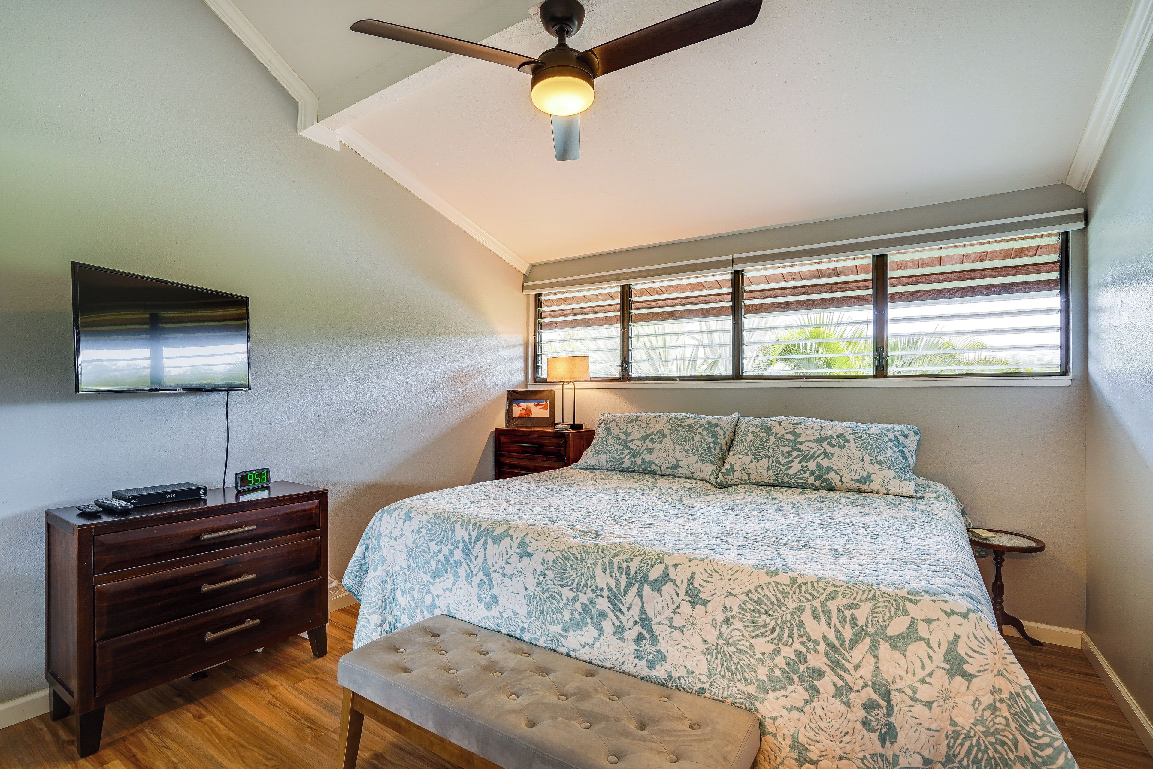 A bedroom with a bed, a TV mounted on the wall, a nightstand, a lamp, and a window with blinds.